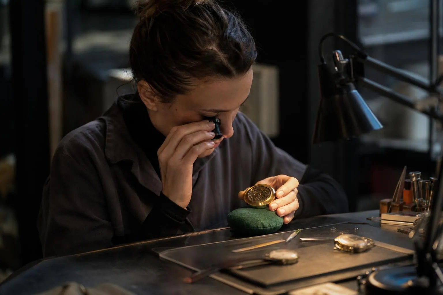 Jeune experte examinant à la loupe le fond d’une montre ancienne posée sur un coussin de velours vert, dans un atelier de bijouterie