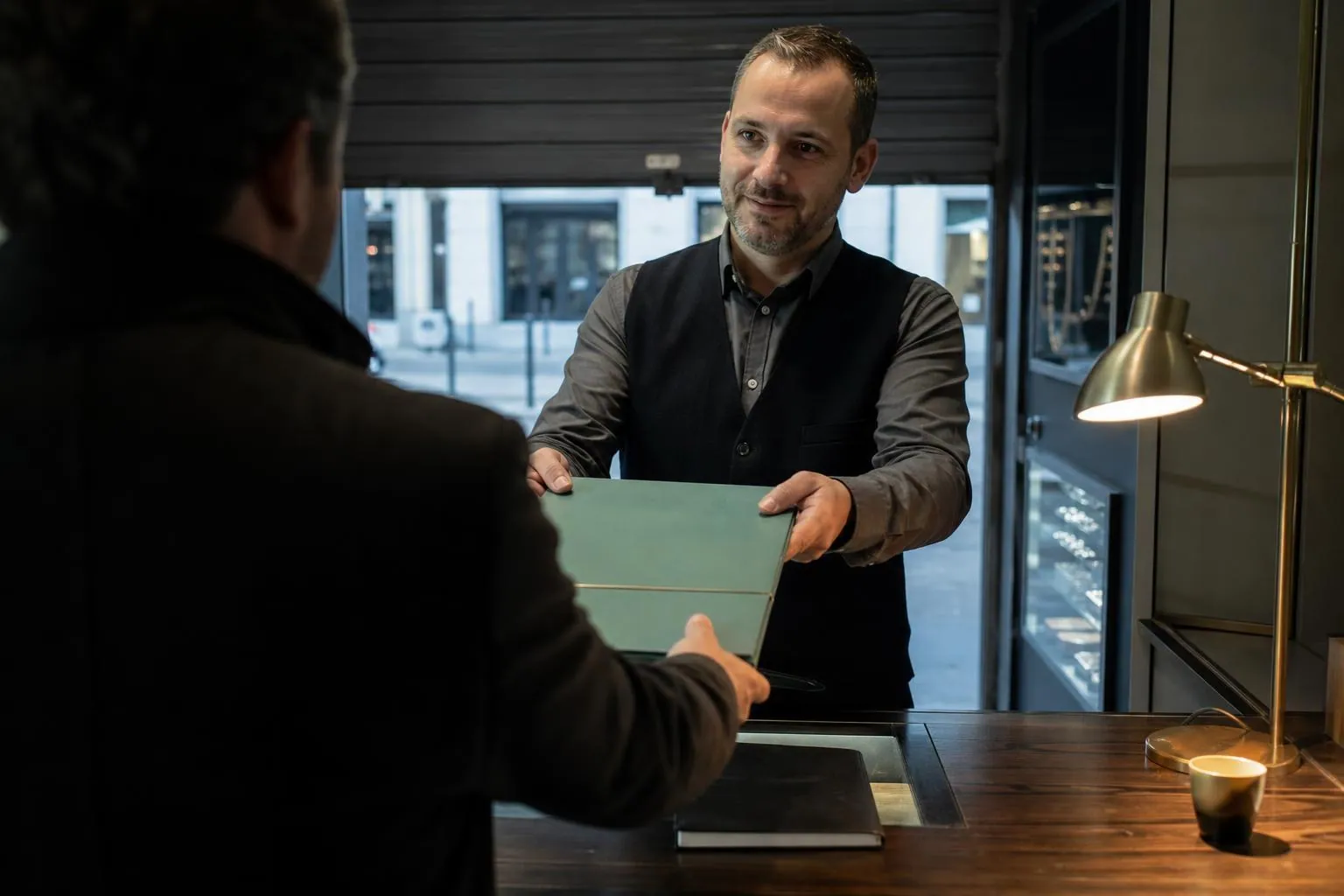 Gérant d’un comptoir tendant une chemise cartonnée fermée par une sangle à un visiteur de dos, devant un rideau métallique à demi relevé